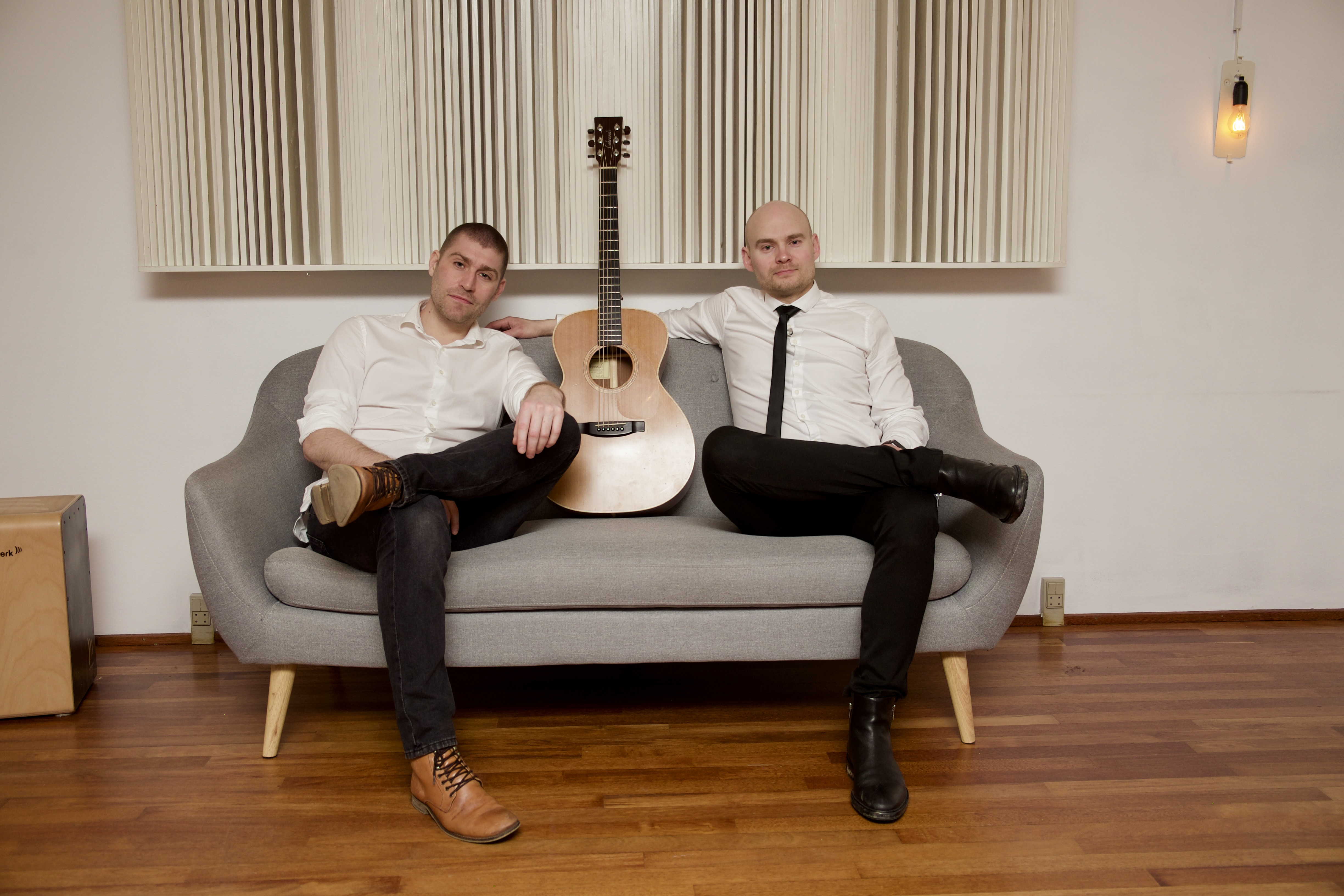 Two men dressed in white shirts and dark pants sit relaxed on a modern gray sofa with a wooden acoustic guitar placed upright between them. The setting is a warmly lit room with wooden floors, minimalist decor, and a soft, inviting atmosphere. The mood is casual and friendly.